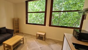 A small, well-lit room with a dark-colored sofa, a wooden table, a small stool, and a window with a view of green trees. - Grand Studio Neuf Climatise Intramuros 9A (Avignon)