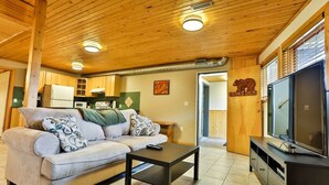 A cozy living room with a sofa, coffee table, and TV. Wooden ceiling and walls, a kitchen in the background, and a bear logo on the wall.
