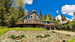 A large, two-story house with a balcony, surrounded by trees and a stone retaining wall.
