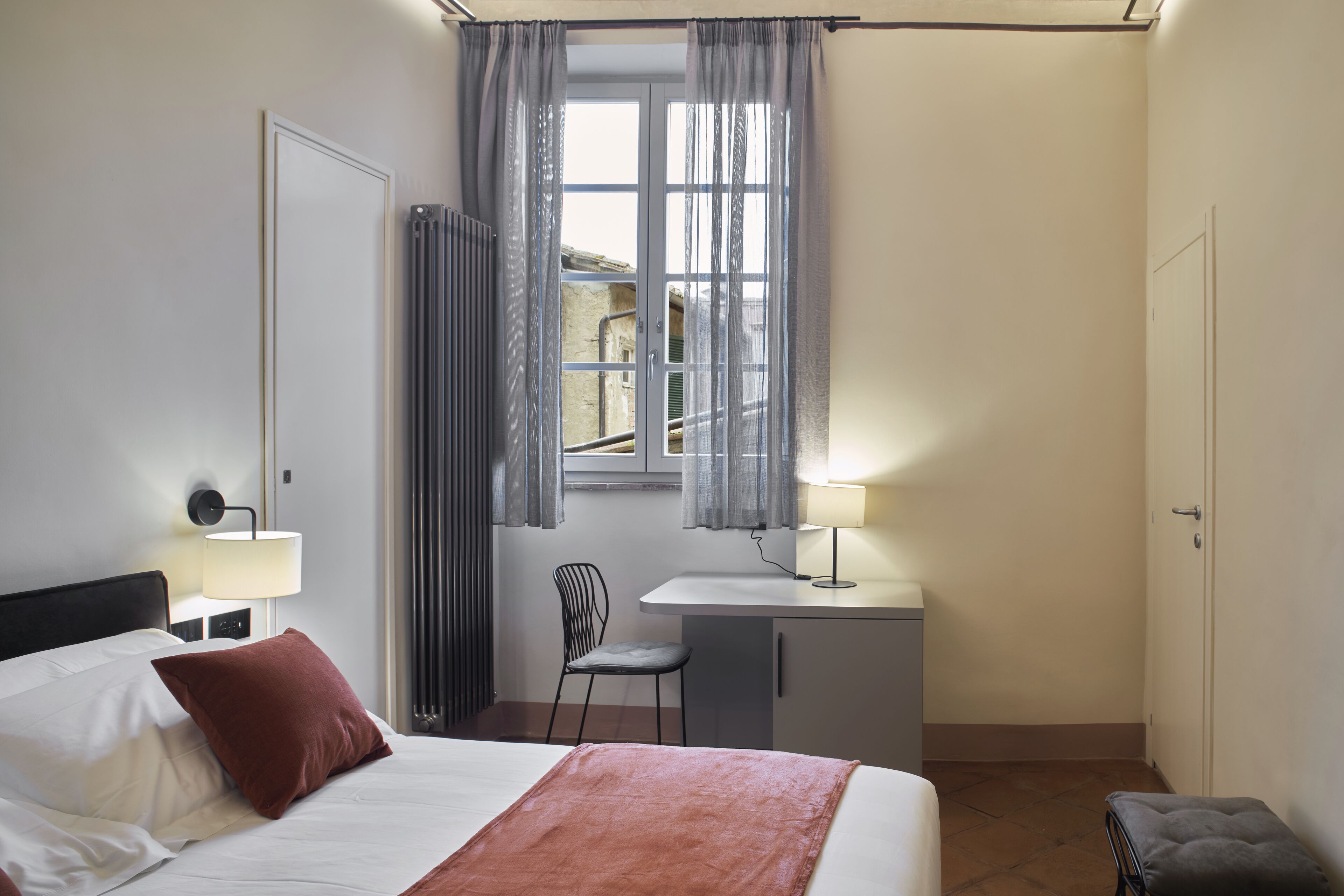 A hotel room with a bed, a desk, a chair, a radiator, and a window with curtains.