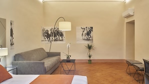 A hotel room with a bed, a sofa, a small table with a lamp, two potted plants, and two wall-mounted air conditioning units.