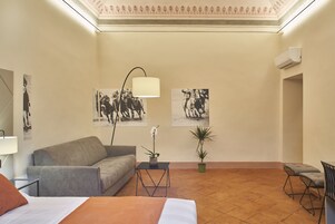 Una camera d'albergo con un letto, un divano, un tavolino con una lampada, due piante in vaso e due condizionatori fissati alla parete.