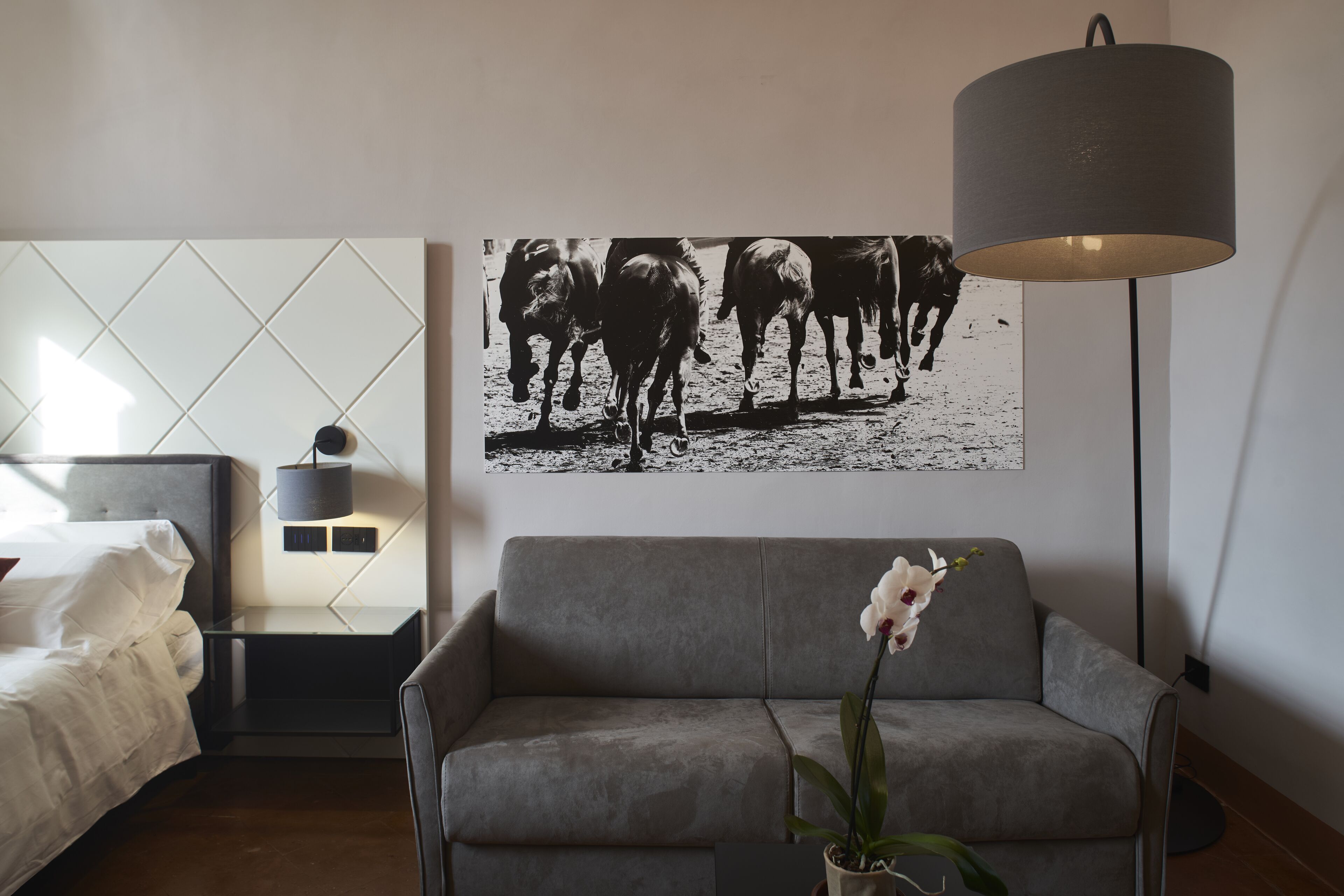 Une chambre moderne avec un lit, un canapé gris, une orchidée en pot, une lampe fixée au mur et une photographie en noir et blanc représentant des chevaux.