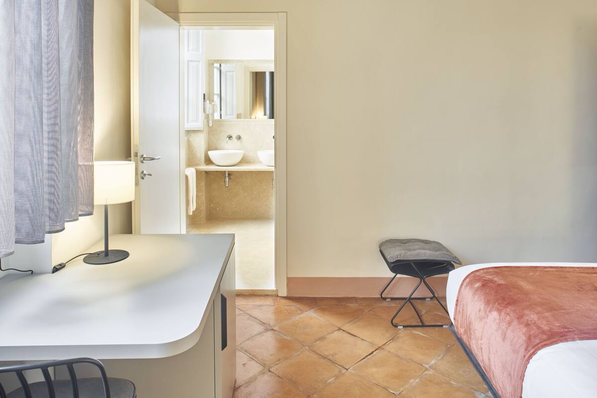 A hotel room with a bed, a desk with a lamp, a chair, a small stool, and a bathroom visible through an open door.