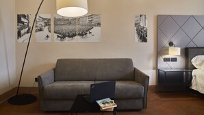 A modern hotel room with a grey sofa, a small table with a laptop and a book, a floor lamp, a bed with a white comforter, a nightstand with a lamp, and black-and-white photographs on the wall.