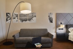 Camera d'albergo moderna con un divano grigio, un tavolino con un portatile e un libro, una lampada da terra, un letto con copriletto bianco, un comodino con una lampada e fotografie in bianco e nero alle pareti.