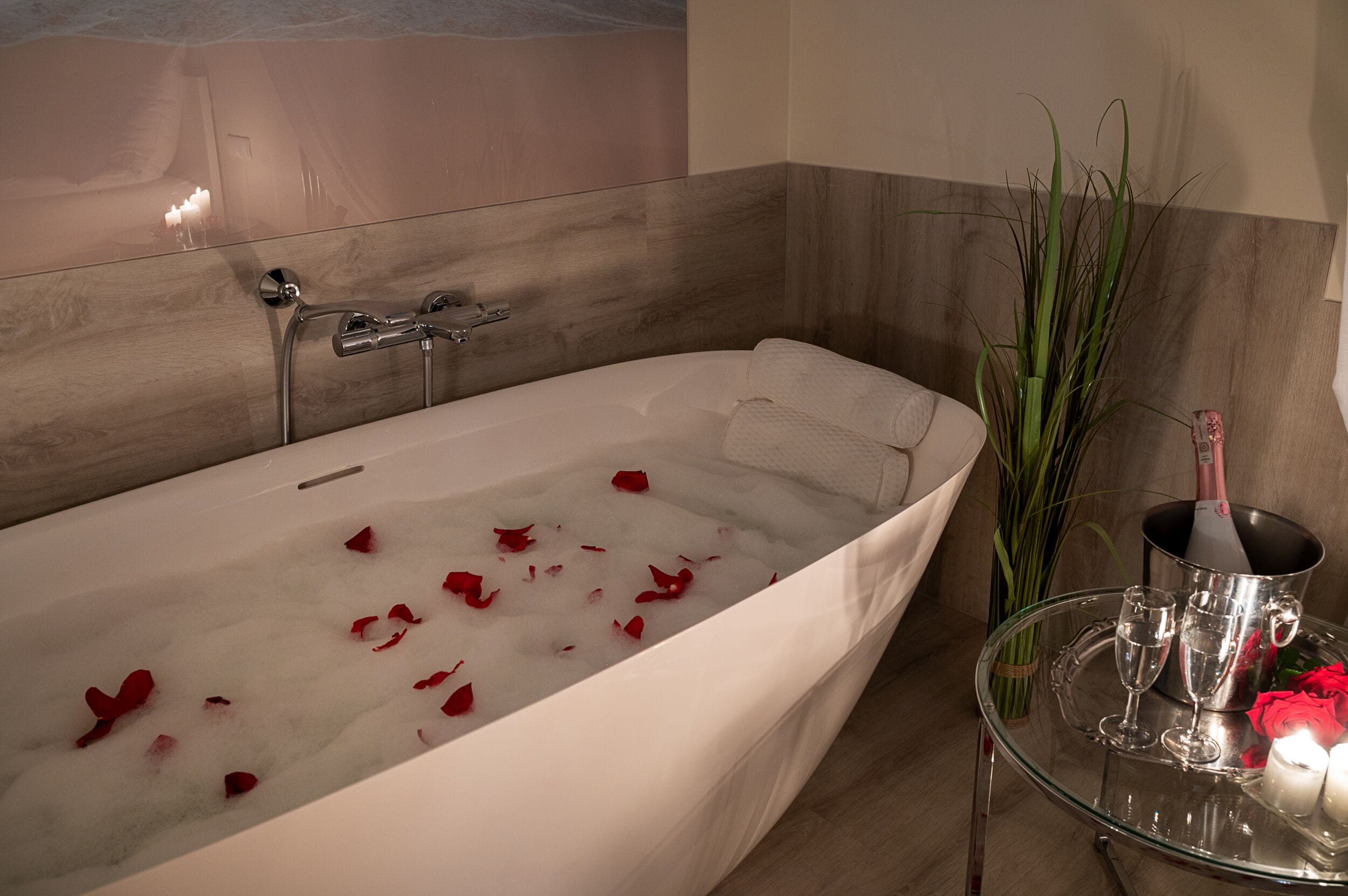 A modern bathroom with a freestanding bathtub, rose petals in the water, a glass table with a bottle of champagne, and a vase with green plants.