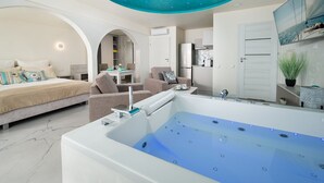 A modern bedroom with a large bed, a sitting area with a sofa and armchairs, a small kitchen area, and a large bathtub with a ceiling light. - Elegante Apartments - Adults only (Wladyslawowo)