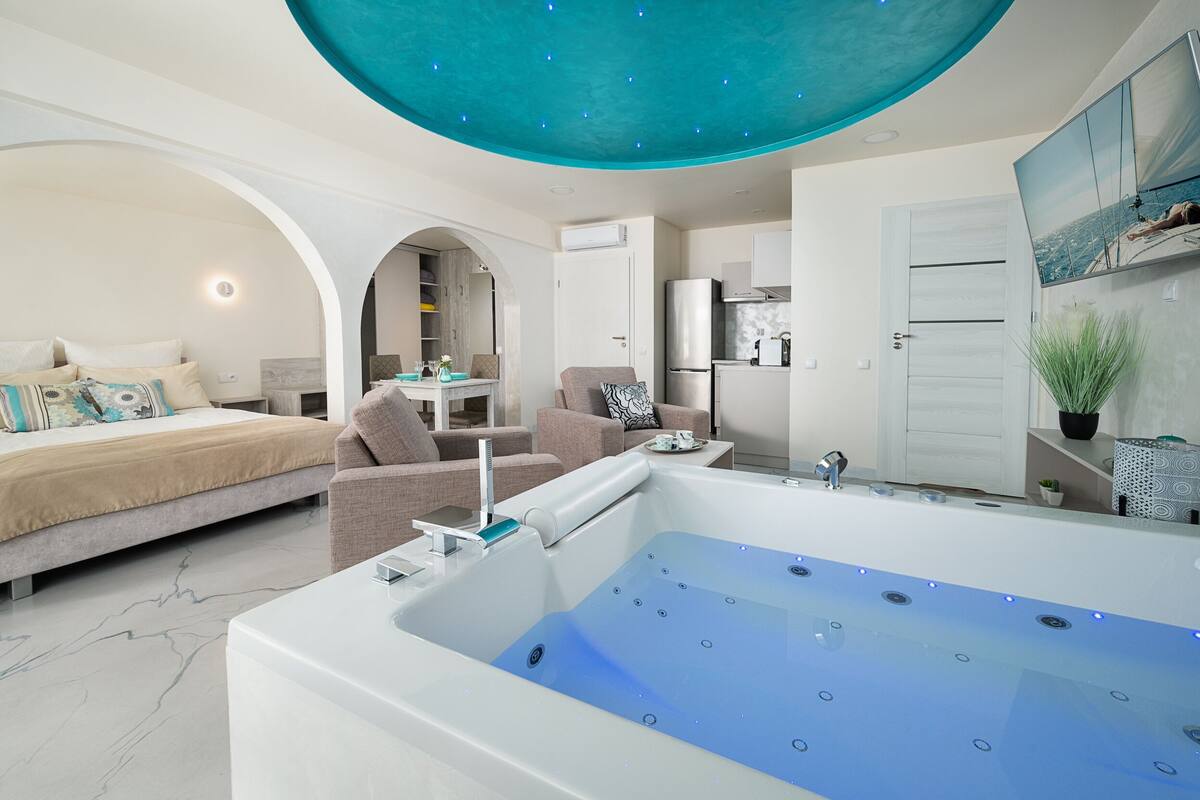 A modern bedroom with a large bed, a sitting area with a sofa and armchairs, a small kitchen area, and a large bathtub with a ceiling light.