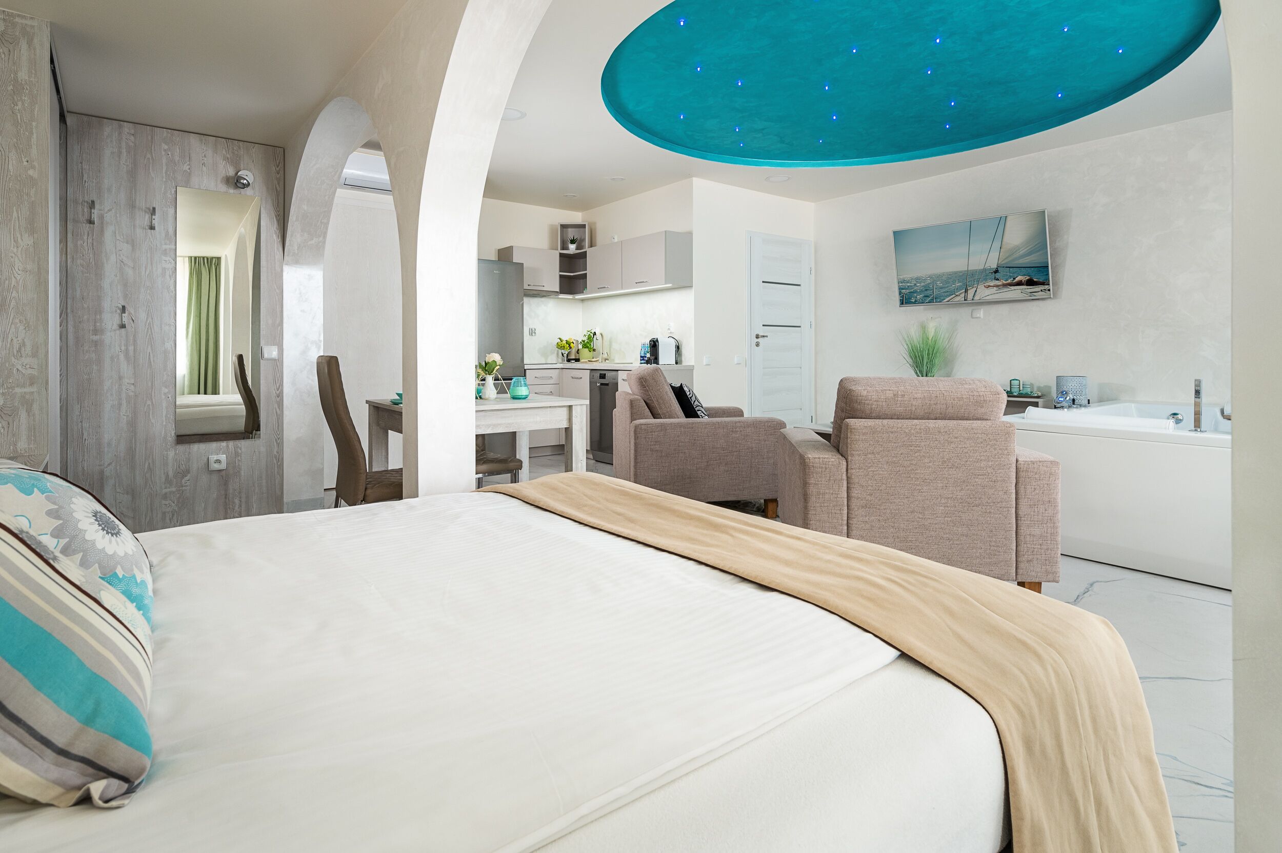 A modern bedroom with a large bed, a seating area with chairs, a kitchen in the background, and a unique ceiling feature.