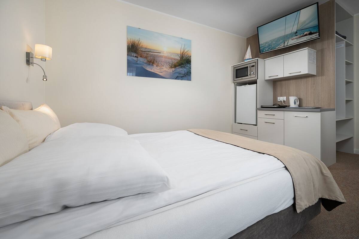 A hotel room with a bed, a flat-screen TV mounted above a wooden cabinet, and a painting of a beach scene on the wall.