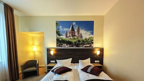 Habitación de hotel con cama doble, dos almohadas, mesita de noche, lámpara, una silla y un ventanal grande con cortinas.