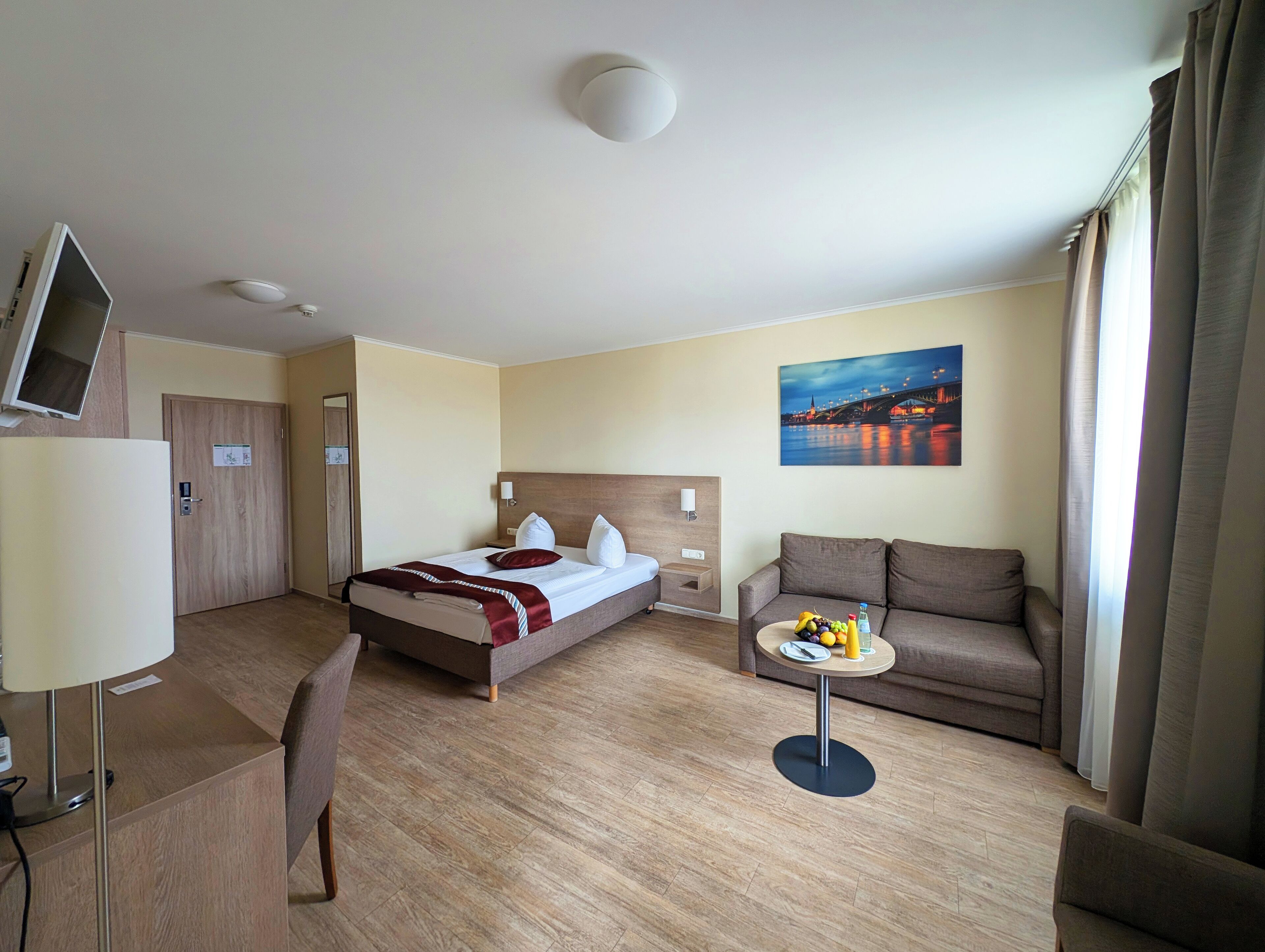 Ein Hotelzimmer mit einem Bett, einem Sofa, einem kleinen Tisch mit Früchten, einem Fernseher und einem Gemälde einer Brücke.