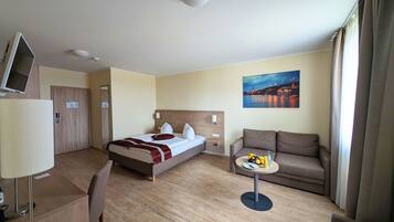 Ein Hotelzimmer mit einem Bett, einem Sofa, einem kleinen Tisch mit Früchten, einem Fernseher und einem Gemälde einer Brücke.
