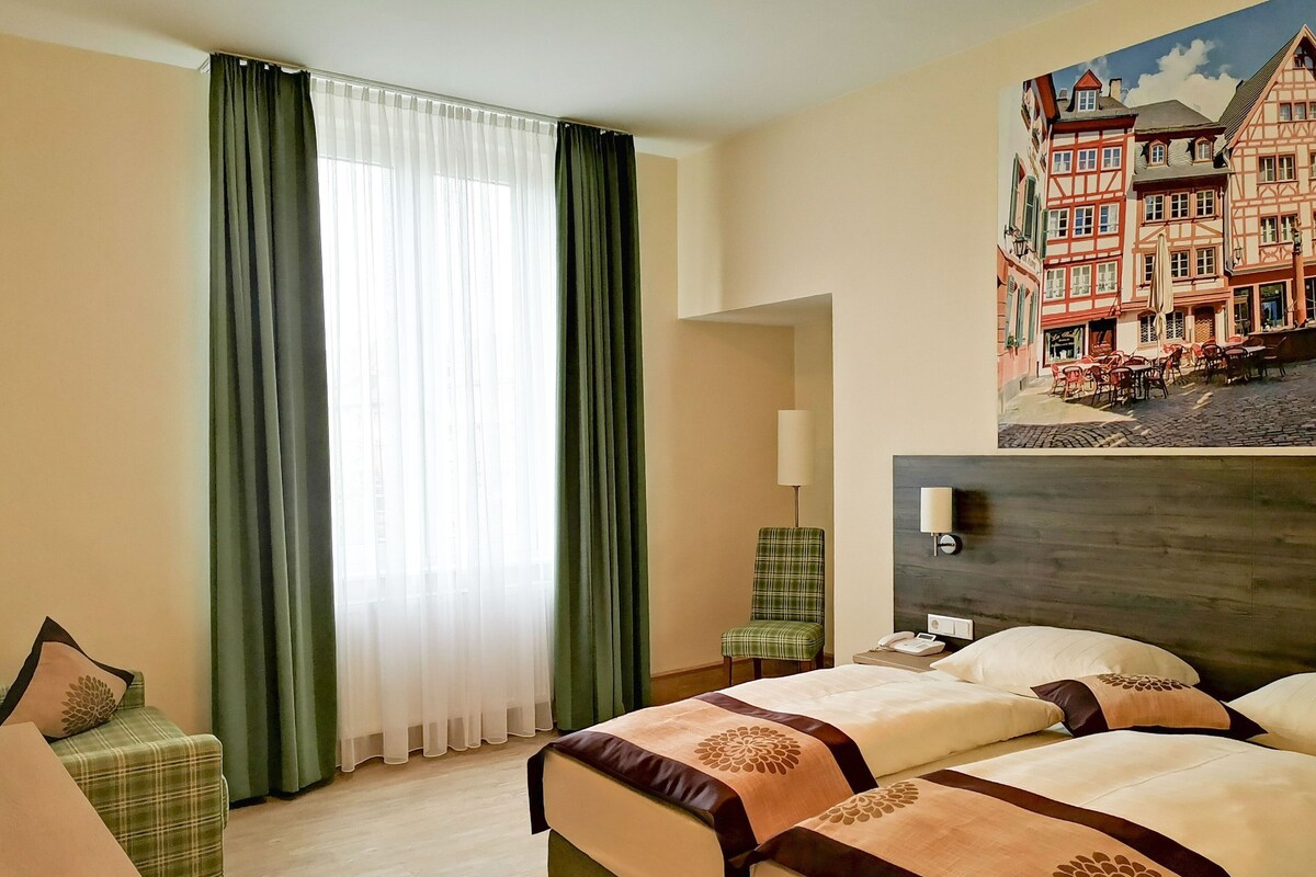A hotel room with two beds, a chair, a lamp, and a painting of a European street scene.