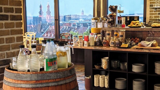 Una barra de desayuno con vista a un paisaje urbano, que incluye un barril de madera, botellas, frascos y una variedad de alimentos para el desayuno.