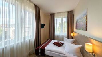 A hotel room with a bed, curtains, a painting, and a TV on the wall.
