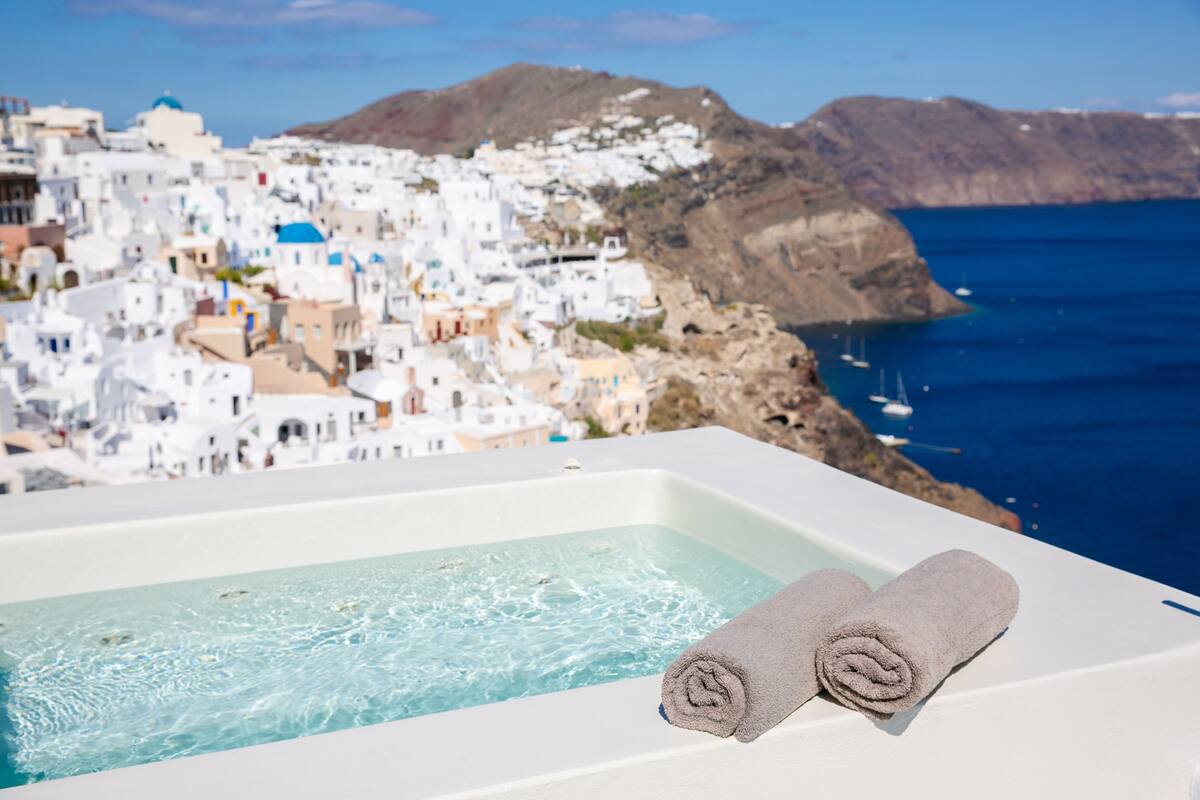 A bathtub with two rolled towels, overlooking a coastal town with white buildings and a view of the sea.