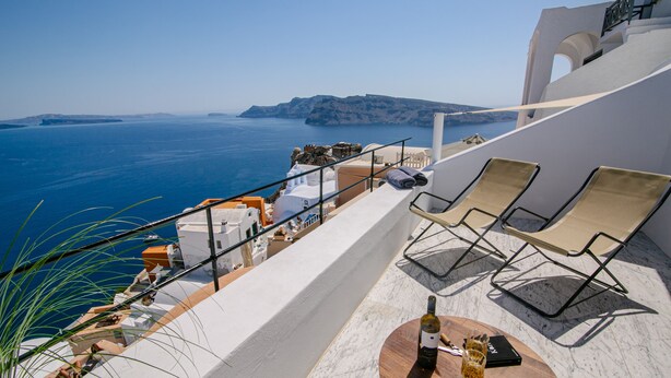 A terrace with a view of the sea, a table with a bottle and glasses, and two lounge chairs. - Platis Residence Villa (Santorini)