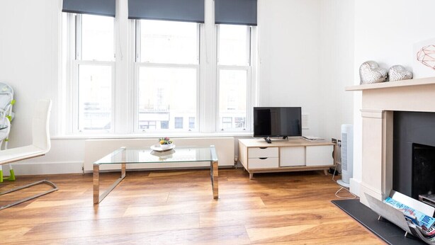 A modern living room with a glass coffee table, a flat-screen TV, a fireplace, and a large window with dark curtains. - Gorgeous Apartment by the Park (SS2) (Greater London)