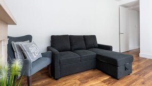 A modern living room with a dark grey sectional sofa, a small armchair, and a decorative pillow with text. The room has wooden flooring and white walls. - Gorgeous Apartment by the Park (SS2) (Greater London)