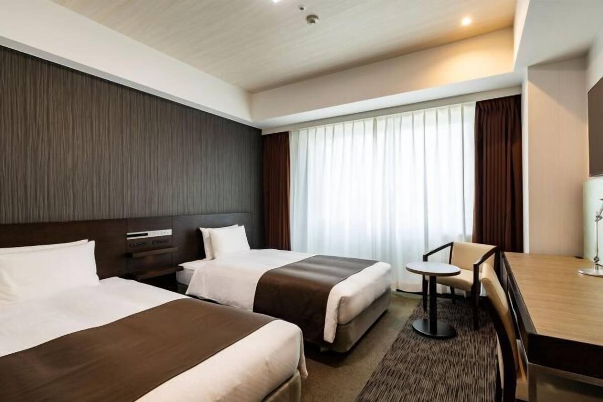 A hotel room with two beds, a desk, a chair, and a large window with curtains.