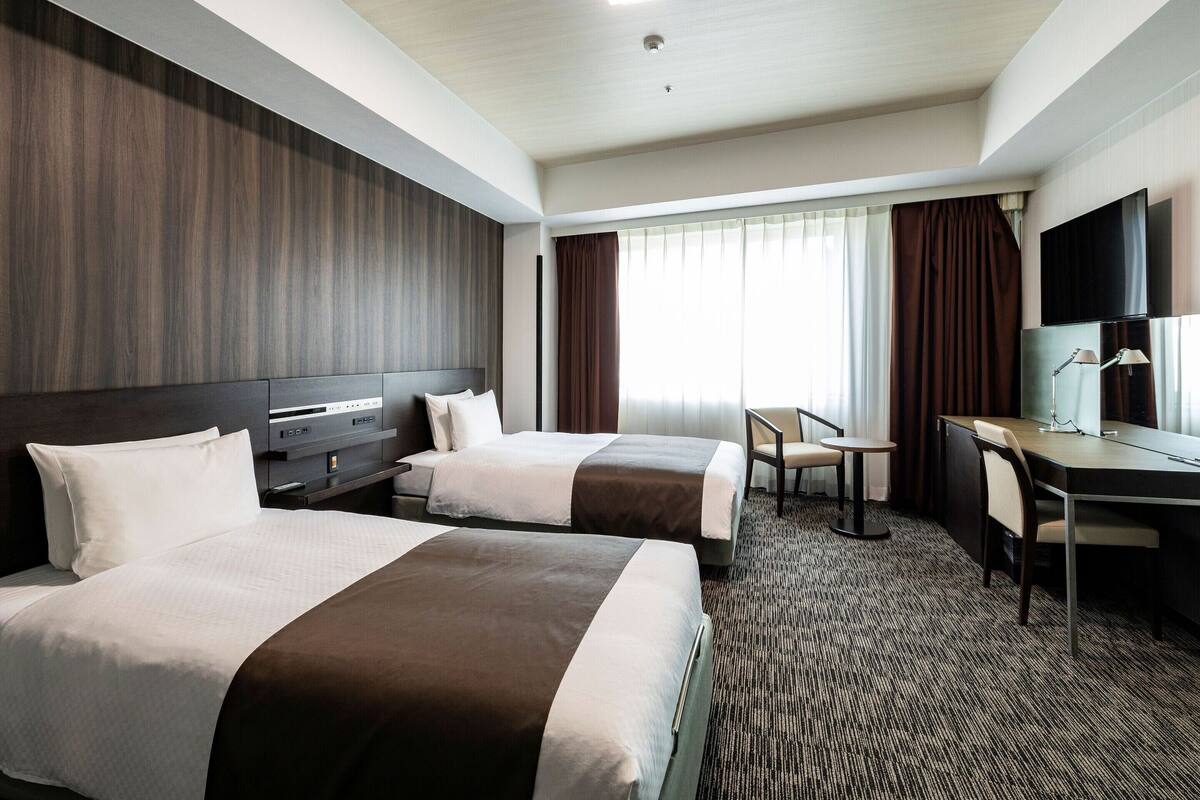 A hotel room with two beds, a desk, a chair, a TV, and a window with curtains.