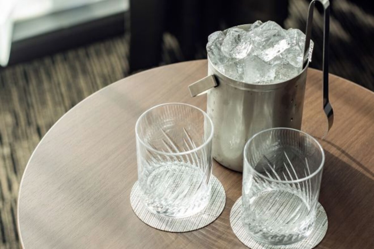 Dos vasos transparentes sobre una mesa redonda, junto a una jarra metálica llena de hielo y con asa.