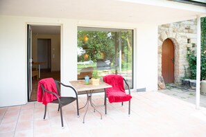 An open patio with a table and two chairs, a wooden door, and a view of a garden through glass doors. - Gartenappartement in der Finca / Spanische Rhön (Hünfeld)