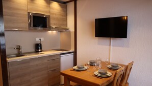 A modern kitchen with wooden cabinets, a built-in microwave, and a small refrigerator. There is a dining table set with plates, glasses, and cutlery. A flat-screen TV is mounted on the wall above the table. - Central Studio at Foot of Les Gets Pistes – Refurbished, Scenic Views, Sleeps 4 (Les Gets)