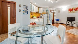 A modern living room with a glass dining table, white chairs, a TV, and a kitchen in the background.