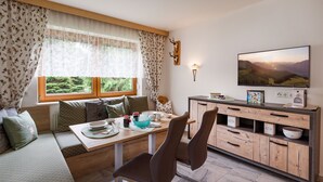 A cozy room with a wooden dining table, two chairs, a sofa with patterned cushions, a television, and a wall-mounted lamp. - Apartment Waltraud (Uderns)