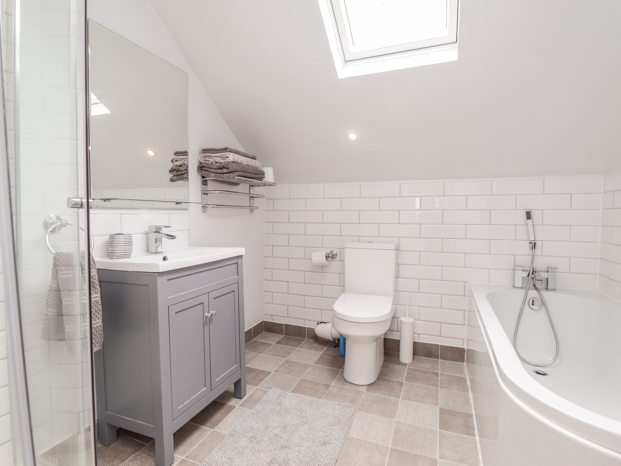 A modern bathroom with a skylight, white tiles, a grey vanity, a toilet, and a bathtub.