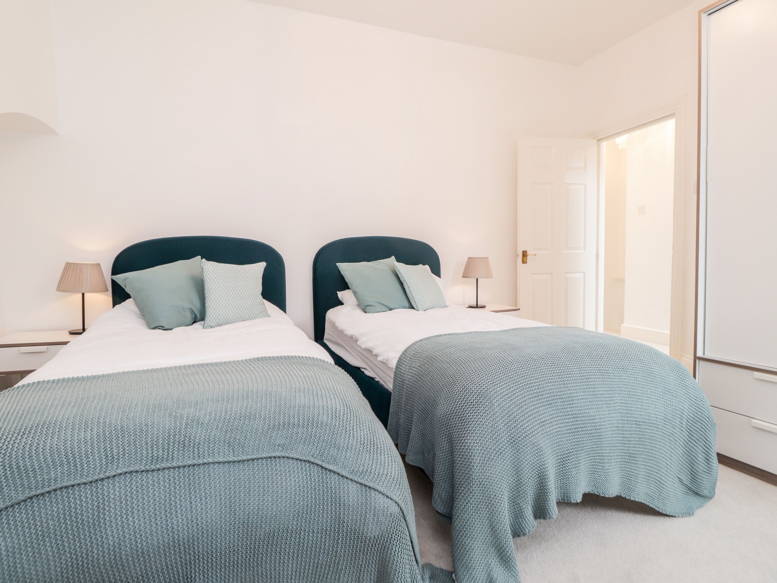 A hotel room with two single beds, each with a teal headboard and matching throw pillows. There are bedside tables with lamps, and a large mirror on the wall.