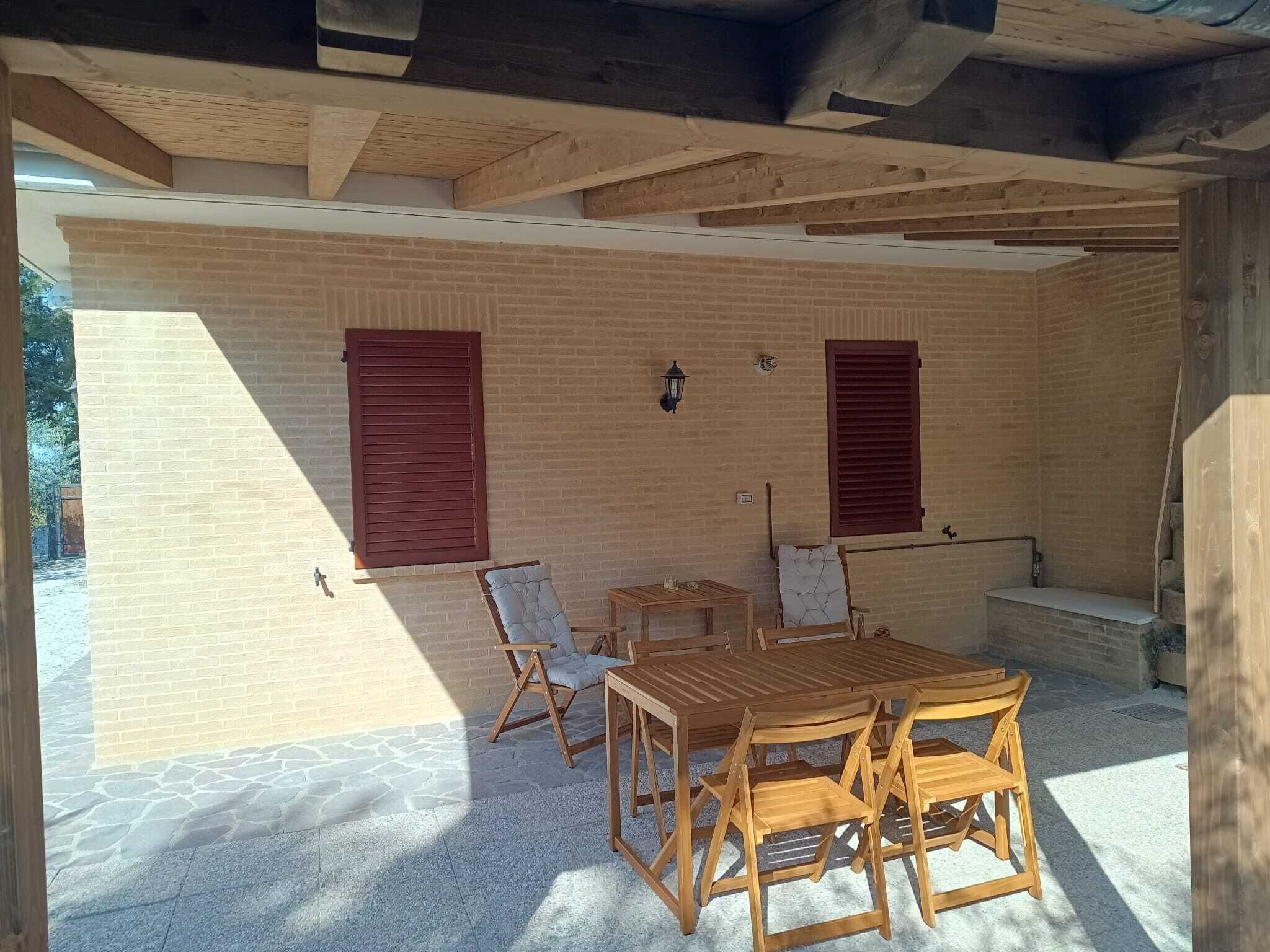 Une terrasse extérieure avec une table et des chaises en bois, un mur en briques et deux fenêtres avec des volets.