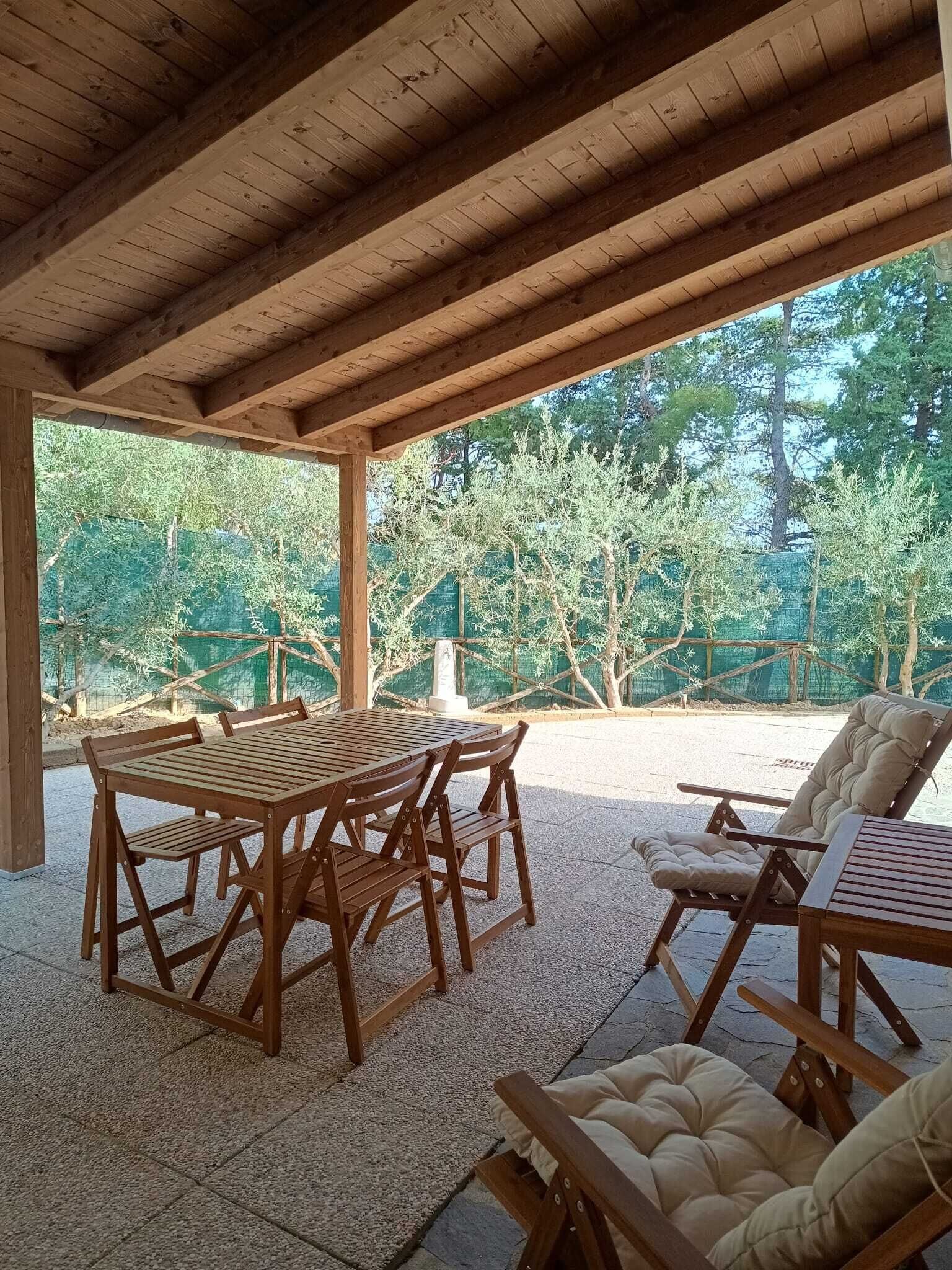 A covered patio with a wooden table and chairs, a cushioned lounge chair, and a view of trees and a fence.