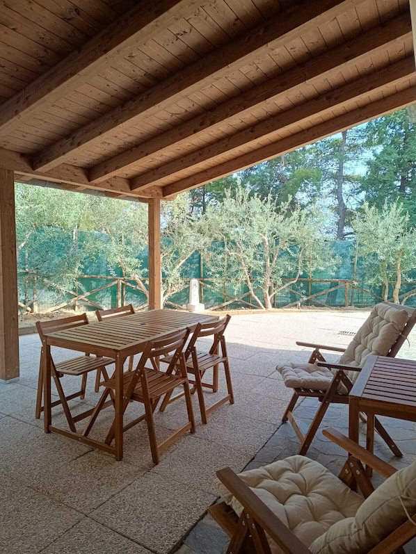 A covered patio with a wooden table and chairs, a cushioned lounge chair, and a view of trees and a fence.