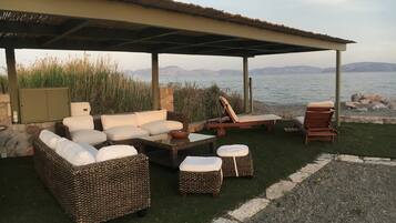 Relax in a serene outdoor seating area surrounded by wicker furniture and a wooden table, with a view of a body of water and distant mountains.