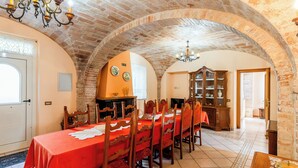 A dining room with a long table, wooden chairs, and a red tablecloth. There is a chandelier, a fireplace, and a cabinet with glass doors.