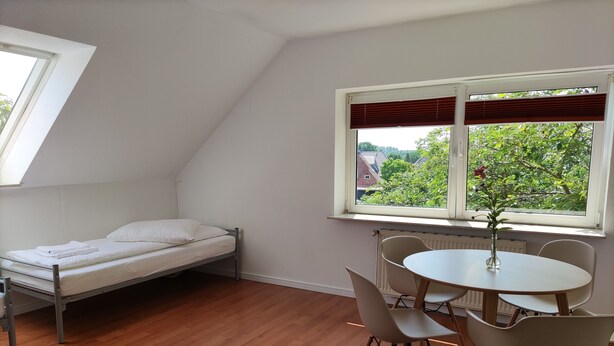 A small, tidy room with a bed, a table with chairs, and a window with blinds. - Ferienwohnung Family Basic in Kiel (Kiel)