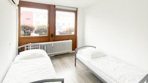 Una habitación pequeña y luminosa con dos camas individuales, un radiador, una ventana y un equipo de aire acondicionado.