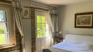 A bedroom with a large bed, a window with curtains, a door, a bedside table with a lamp, and a framed picture on the wall.