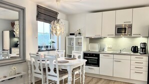 A modern kitchen with white cabinetry, a dining table with chairs, a chandelier, and a view of the outdoors. - fewo1846 - MuschelNest (App. (Harrislee)