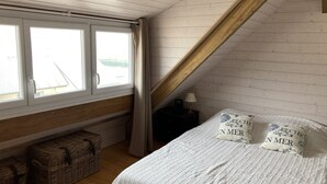 Une chambre avec un plafond en pente, un lit avec une literie blanche, deux oreillers ornés de motifs, une table de chevet avec une lampe et une fenêtre avec des rideaux.