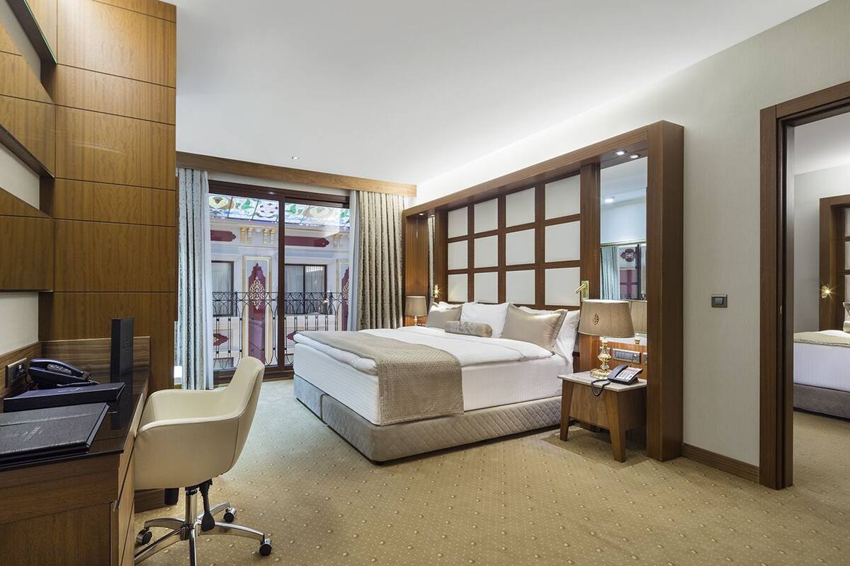 A hotel room with a large bed, a desk with a chair, a bedside table with a lamp, and a mirror.