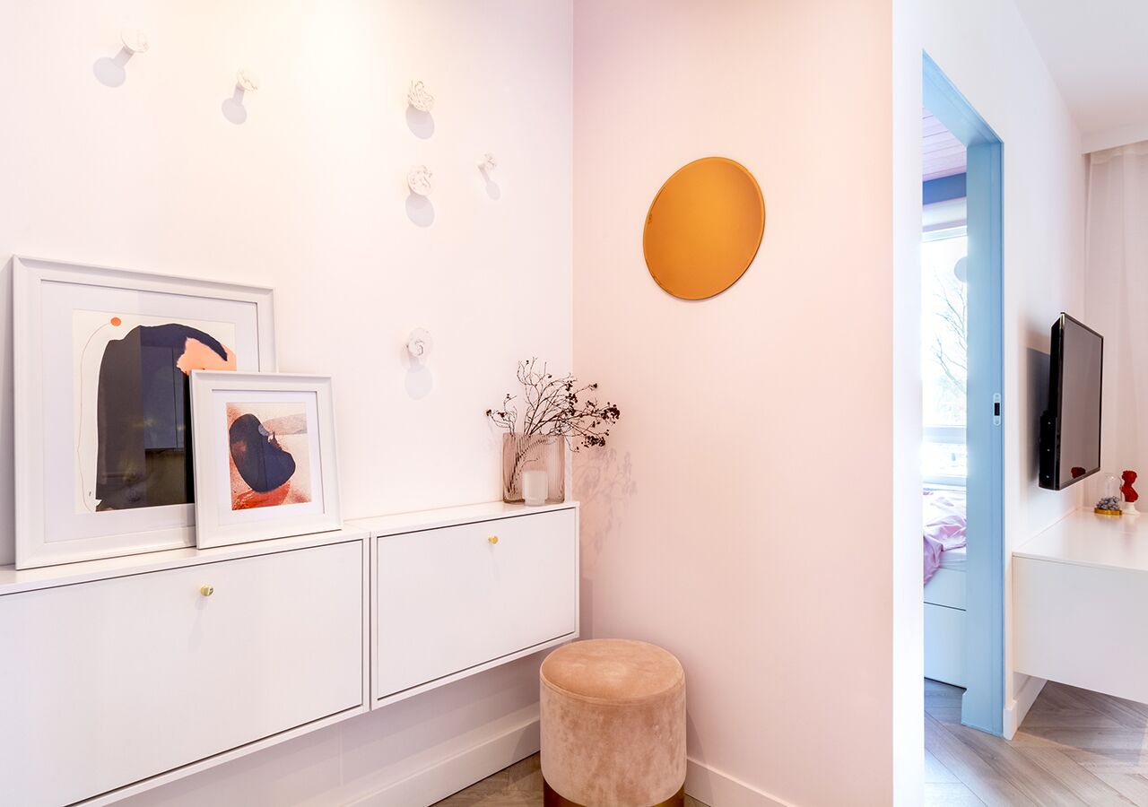 Un intérieur moderne avec une console blanche, des œuvres d’art encadrées, un miroir rond et un petit tabouret rond.