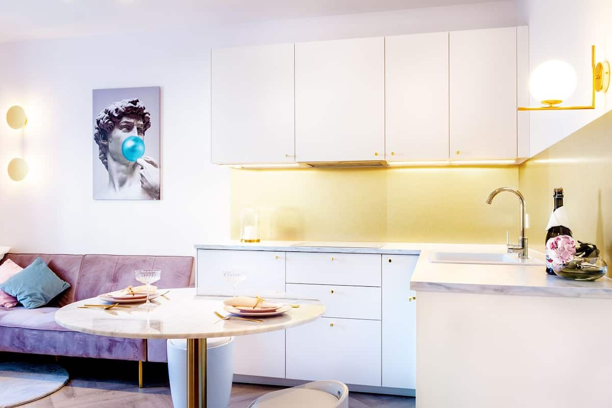 Una cocina moderna con armarios blancos, encimera de mármol y un lavamanos. Hay una mesa de comedor con platos y una copa.