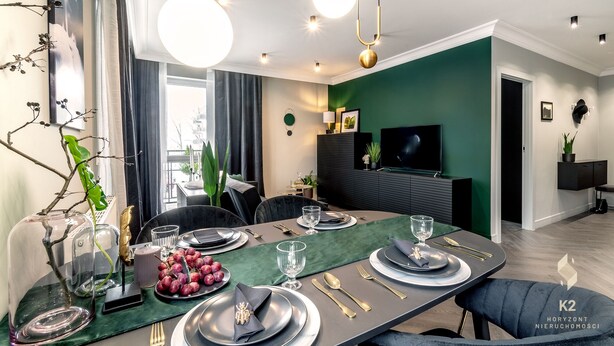 Une salle à manger moderne avec un mur d’accent vert foncé, une table ronde dressée pour quatre personnes et un téléviseur fixé au mur.
