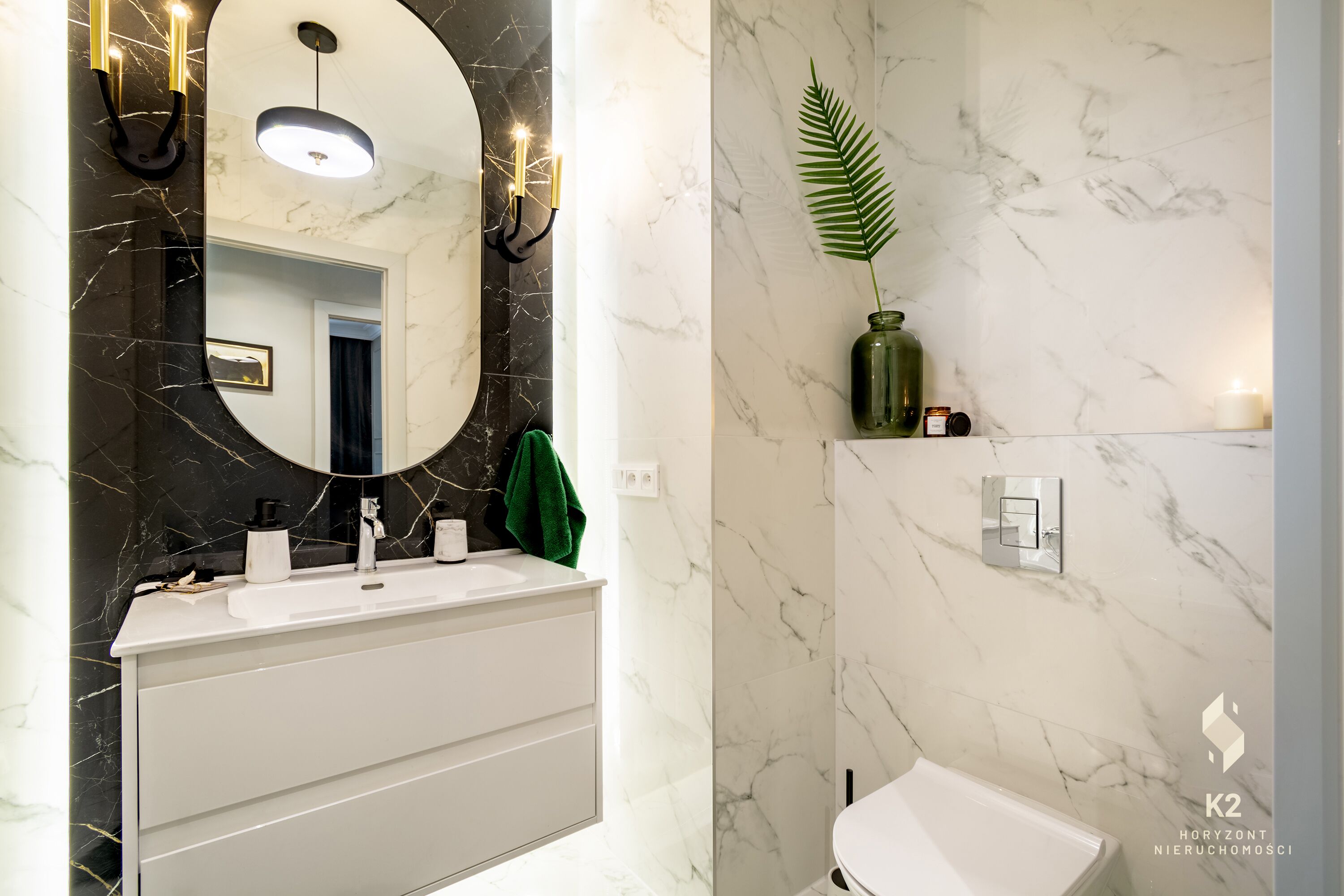 A modern bathroom with a white vanity, a large mirror with a circular frame, a green plant, and a white toilet.