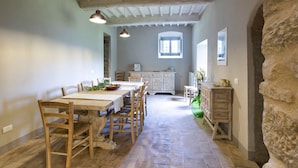 Comedor rústico con mesa y sillas de madera, pared de piedra y vista a una cocina al fondo.